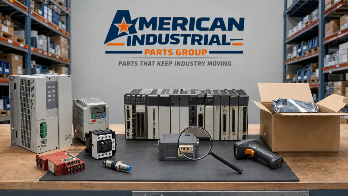 A collection of industrial electronic components, including PLC relays, a motor drive, a contactor, a magnifying glass, and a handheld tool, arranged on a workbench in front of a gray wall with the 'American Industrial Parts Group' logo. Shelves filled with boxes are visible in the background.